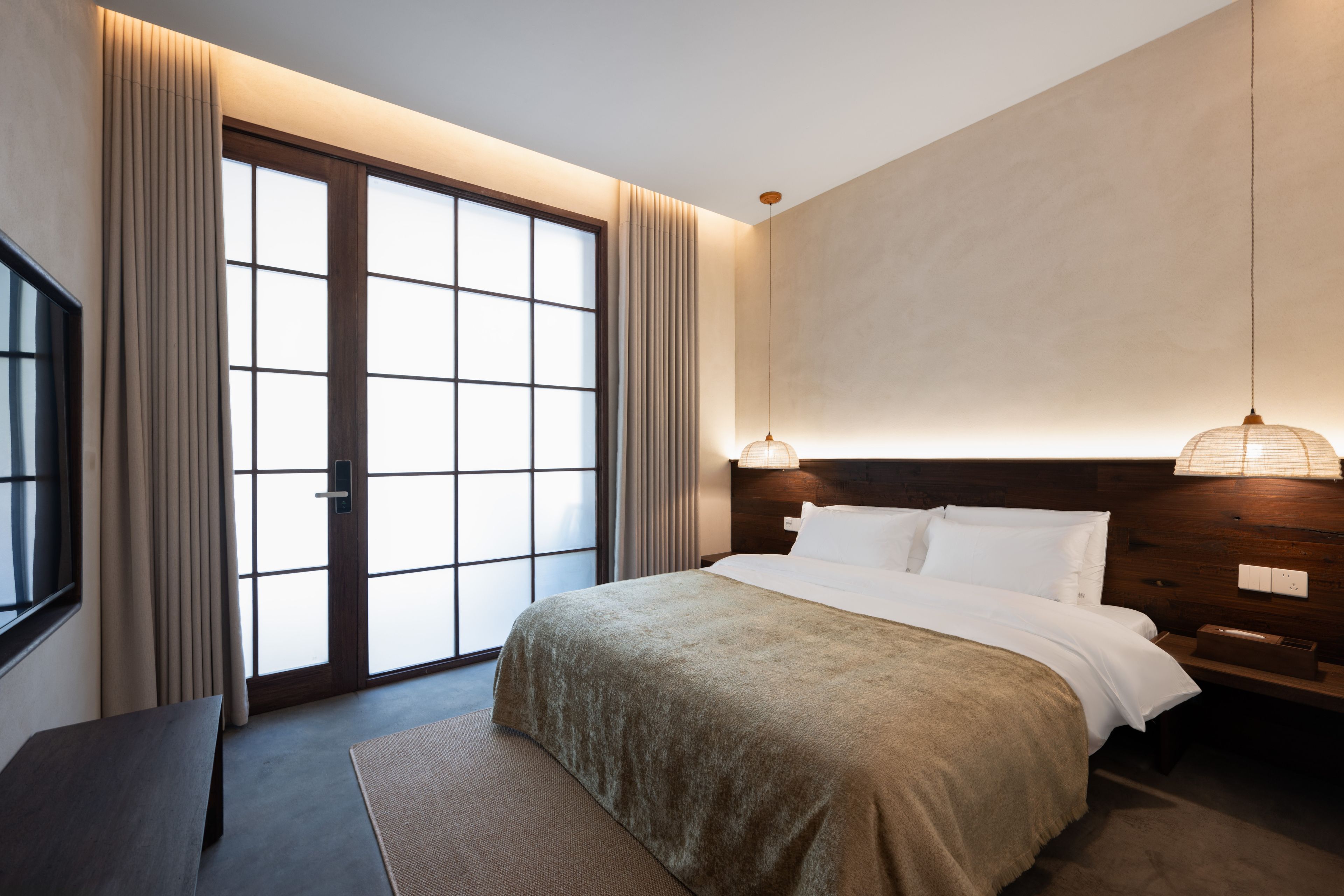 A modern hotel room with a large bed, a bedside table, a desk, and a view through a sliding door.