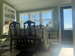 A dining area with a table set for four, flanked by chairs with floral upholstery. A bookshelf is visible to the left, and a window with a view of trees is on the right.