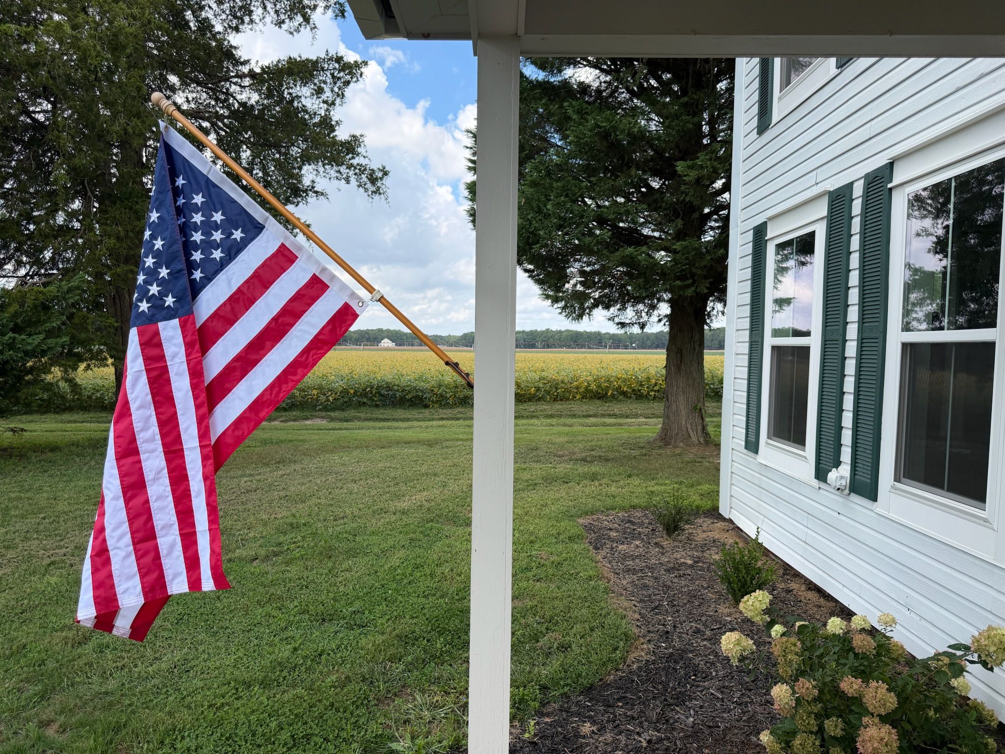 A residential property with a flagpole, a house with green shutters, and a field in the background.