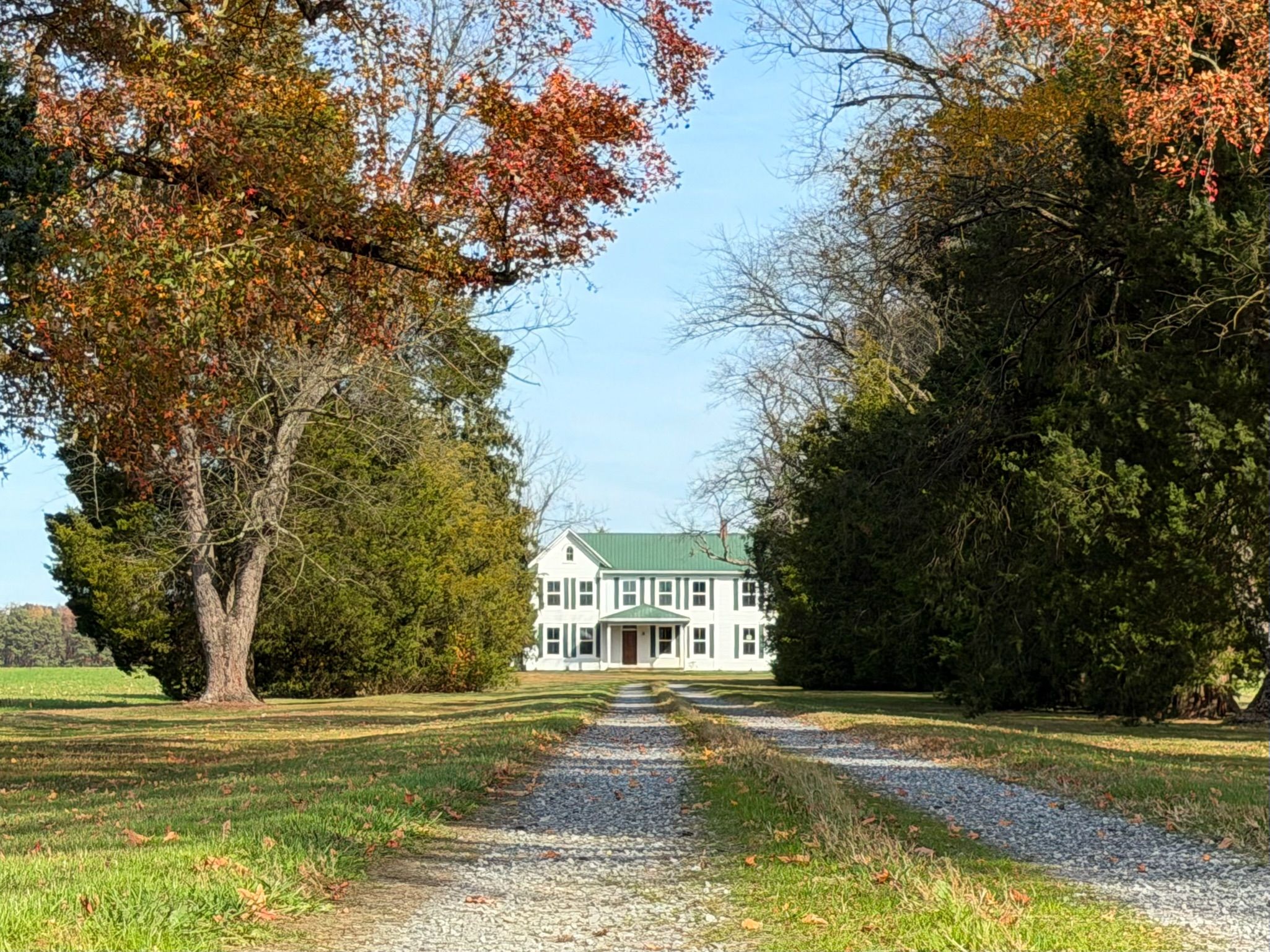 A two-story white building with a green roof, surrounded by trees and a gravel path leading to it.