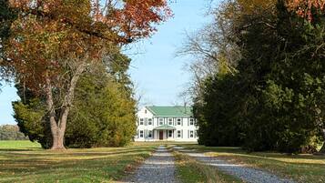 A two-story white building with a green roof, surrounded by trees and a gravel path leading to it.