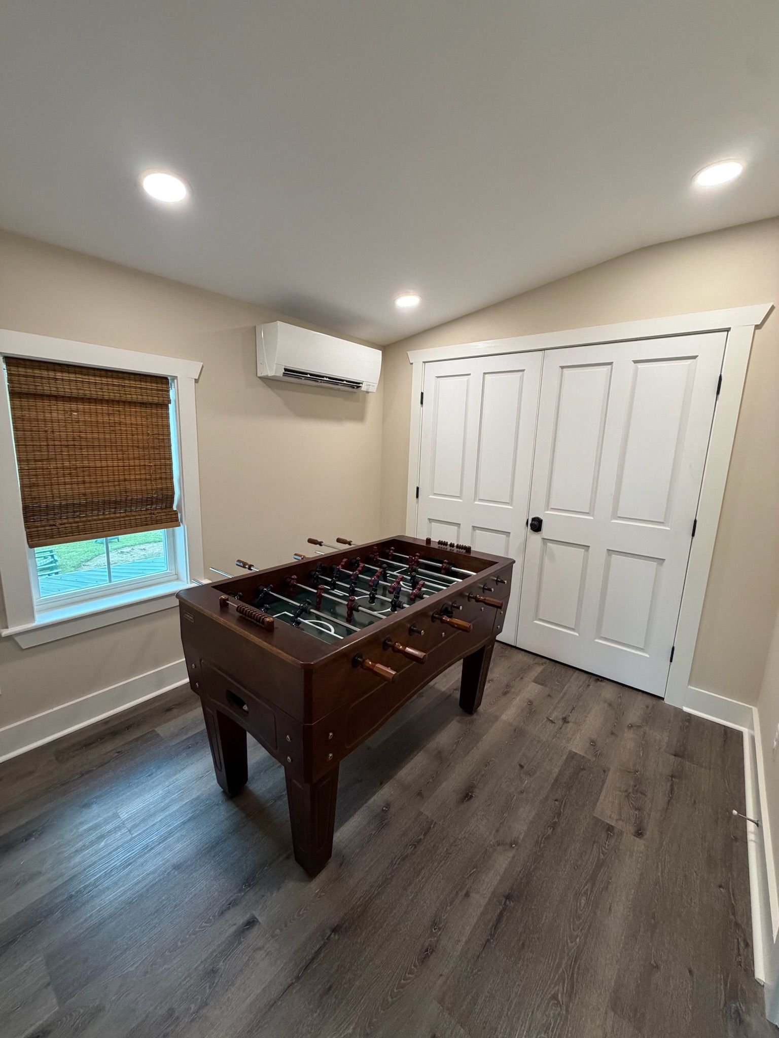 Una habitación con mesa de futbolín de madera, una ventana con persianas, una puerta y un equipo de aire acondicionado en la pared.