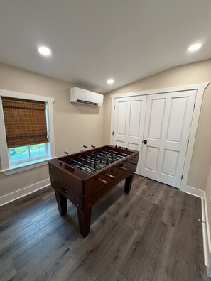 A room with a wooden foosball table, a window with blinds, a door, and an air conditioning unit on the wall.