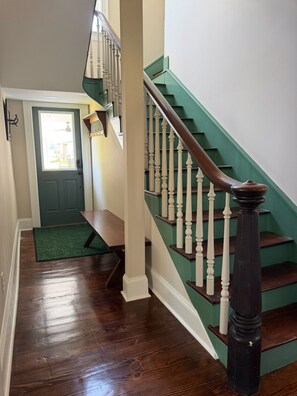 Una escalera con barandilla de madera y balaustres blancos, que lleva a un rellano con un banco y una puerta.