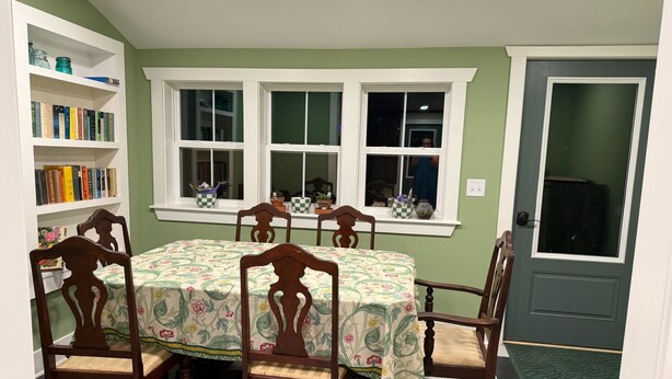 Uma sala de jantar com uma mesa posta para seis pessoas, rodeada por cadeiras de madeira, uma estante de livros e uma janela com plantas em vasos.