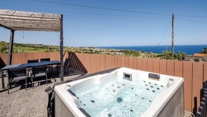 A hot tub with a wooden deck and outdoor seating area, overlooking a coastal landscape. - Northeast Wind | Basalt Lodge with AC & Jacuzzi and Ocean Views (Lomba da Fazenda)