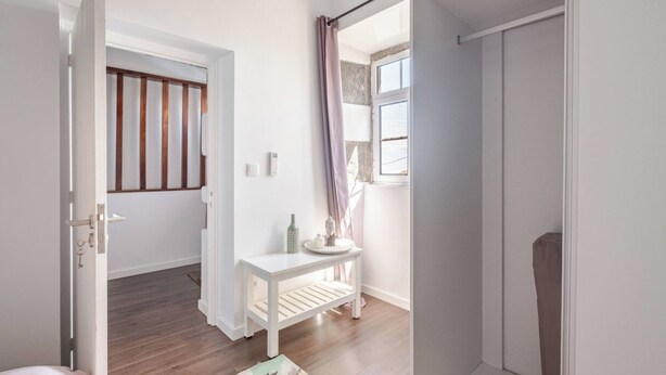A room with a wooden floor, a white door, a small white table with a vase and a bottle, and a window with a curtain. - Northeast Wind | Basalt Lodge with AC & Jacuzzi and Ocean Views (Lomba da Fazenda)