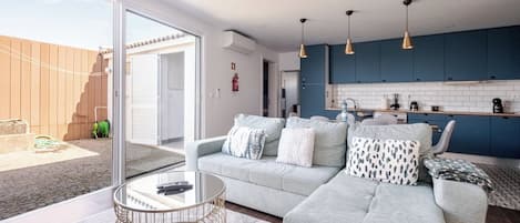 A modern living room with a grey sofa, a glass coffee table, and a kitchen area with blue cabinets and pendant lights.