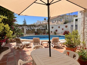 A poolside area with a table and chairs, surrounded by potted plants and a view of a building with a tiled roof. - Apartment in Nerja Near Burriana Beach (Nerja)