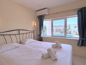 A hotel room with a bed, an air conditioning unit, a window with curtains, and a view of a pool and plants outside. - Apartment in Nerja Near Burriana Beach (Nerja)