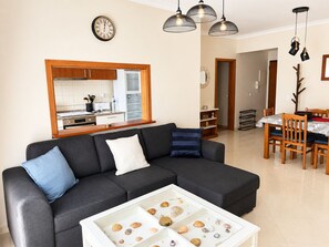 A modern living room with a sectional sofa, a coffee table with seashells, a dining area with wooden chairs, and a kitchen in the background. - Dolly Monte Verde - Apartment with Ocean View (Faro)