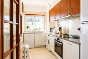 A compact kitchen with white appliances, wooden cabinets, and a window with a view. - Dolly Monte Verde - Apartment with Ocean View (Faro)