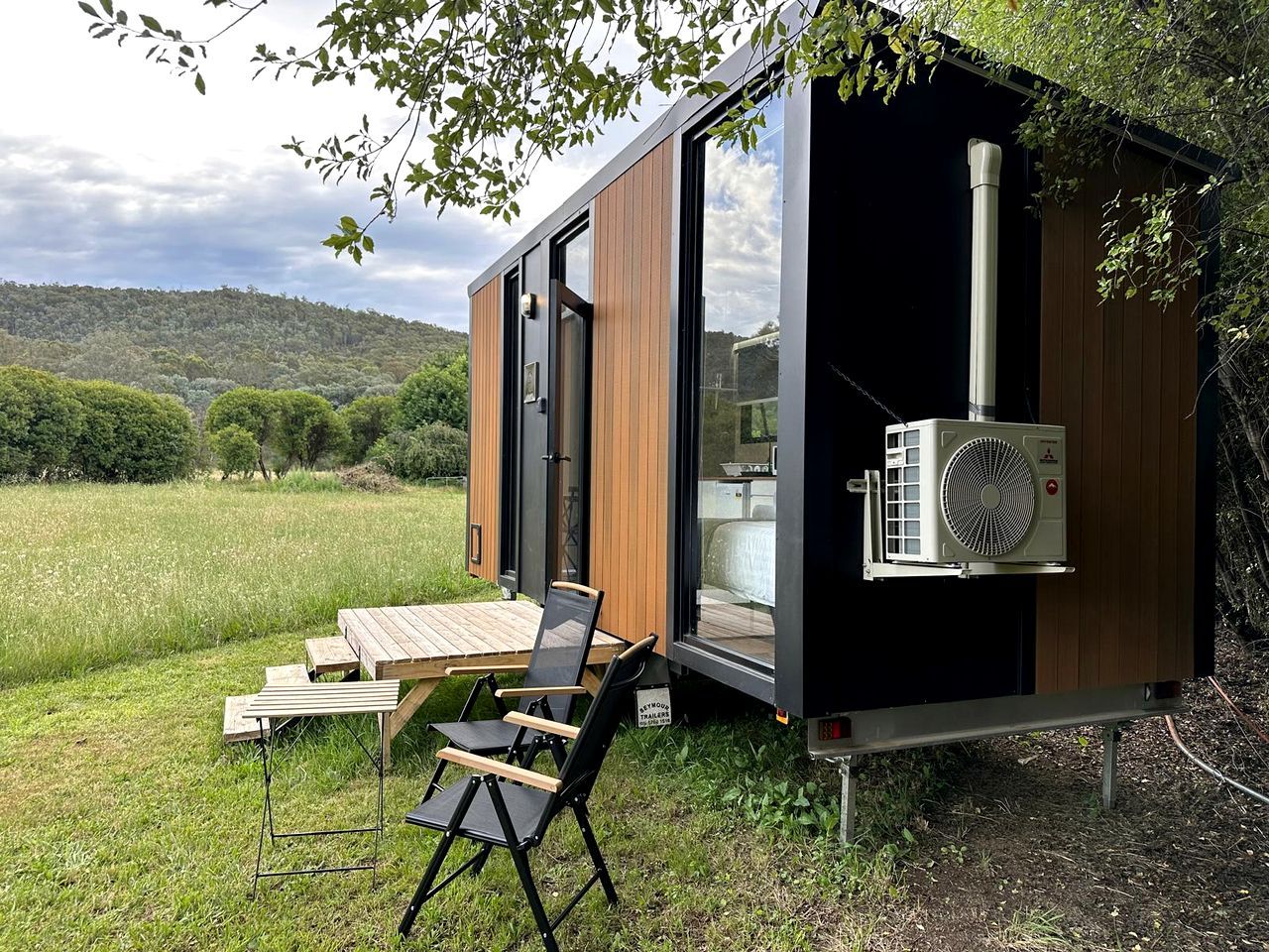 A modern, compact mobile home with a sliding door, a small deck with a table and chairs, and an air conditioning unit.