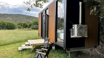 A modern, compact mobile home with a sliding door, a small deck with a table and chairs, and an air conditioning unit.