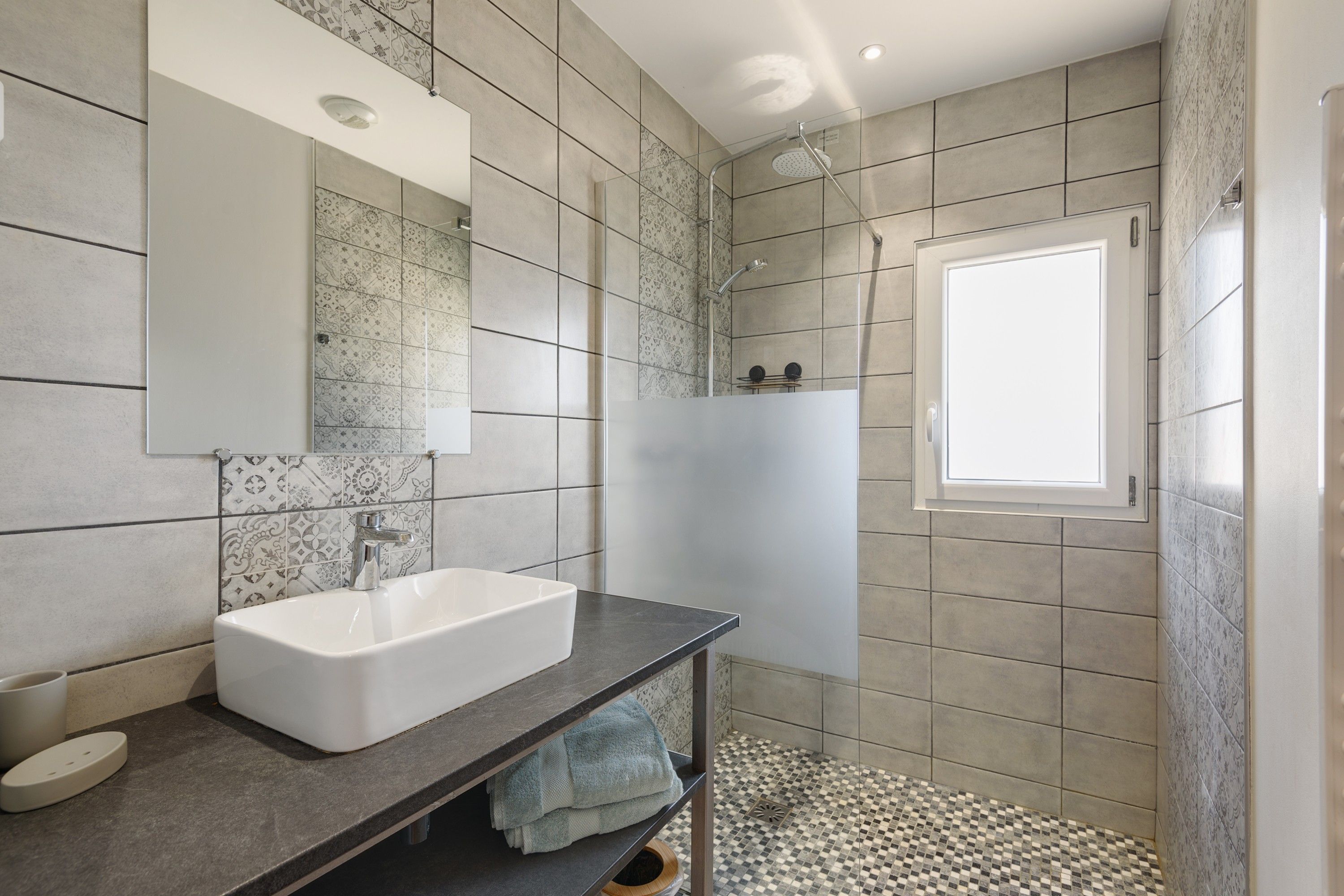 A modern bathroom with a white sink, dark countertop, patterned tiles, a shower with a glass door, and a small window.