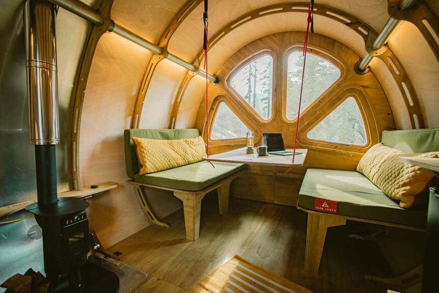 A cozy interior with a curved wooden structure, two green cushioned seating areas, a table with a laptop, and a wood-burning stove.