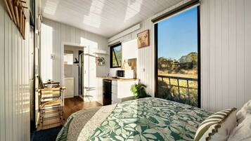 A bedroom with a large bed, a view of the outdoors, a kitchenette, and a door leading to another room.