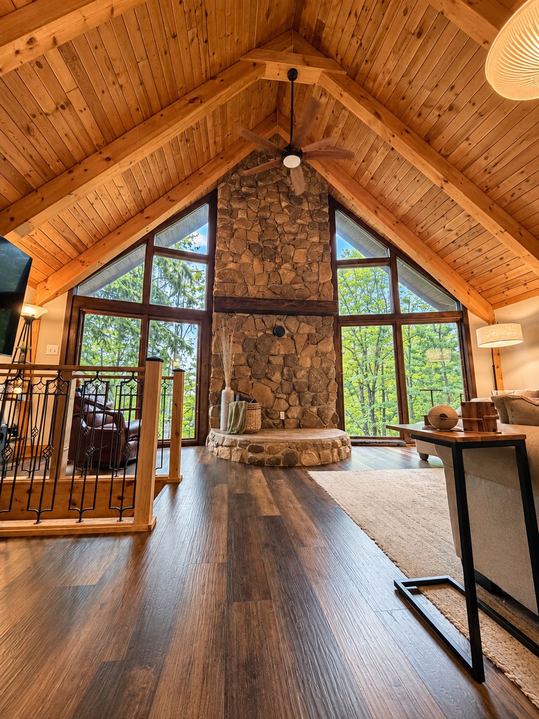 A spacious room with a high ceiling, a stone fireplace, and large windows offering a view of the outdoors.