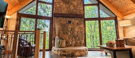 A spacious room with a high ceiling, a stone fireplace, and large windows offering a view of the outdoors.