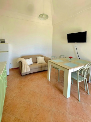 Ein Zimmer mit einer Couch, einem Esstisch mit Stühlen, einem an der Wand montierten Flachbildfernseher und einem gefliesten Boden.