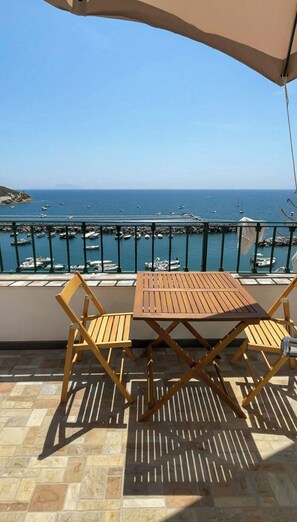 Uma varanda com mesa e cadeiras de madeira, com vista para uma marina com barcos e um céu azul límpido.