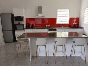 A modern kitchen with a central island, bar stools, a refrigerator, and a window with blinds. - Villa Indy (Sainte-Anne)