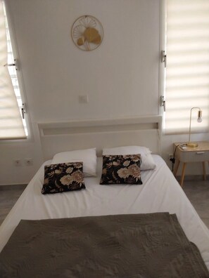 A bed with white bedding and two floral pillows, a bedside table with a lamp, and a wall-mounted light fixture. - Villa Indy (Sainte-Anne)