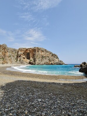 Ein felsiger Strand mit klarem, blauem Wasser, ein Kiesstreifen im Vordergrund und eine zerklüftete Klippe im Hintergrund.