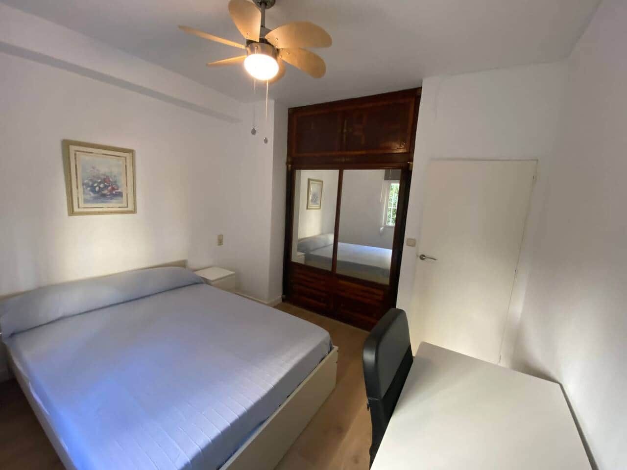 Une chambre à coucher comprenant un lit, un bureau, un ventilateur de plafond et une porte donnant sur une autre pièce.
