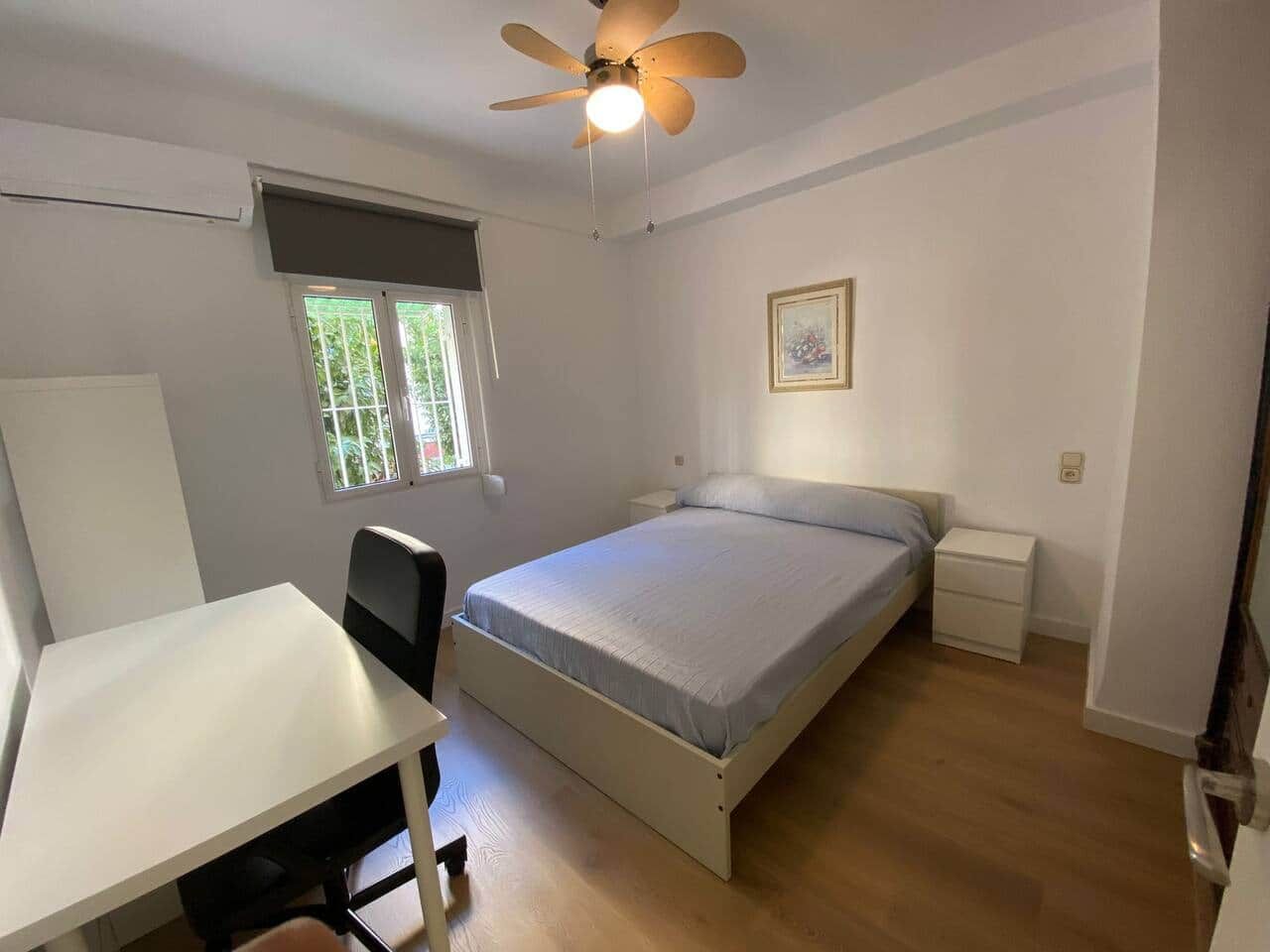Ein Schlafzimmer mit Bett, Schreibtisch, Stuhl, Deckenventilator, Fenster und Nachttisch.