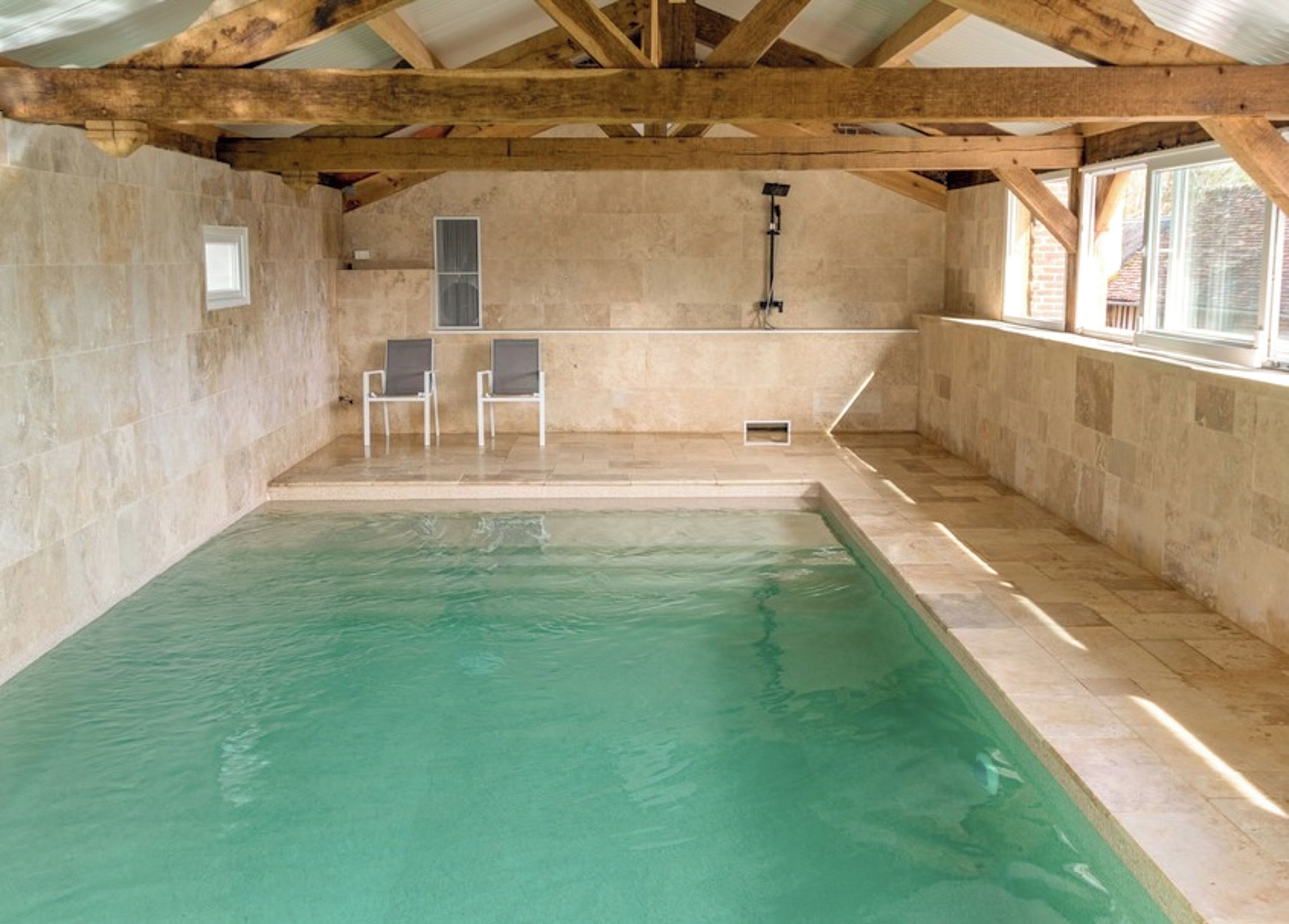 Une piscine intérieure avec un mur en pierre, des poutres en bois et deux chaises.