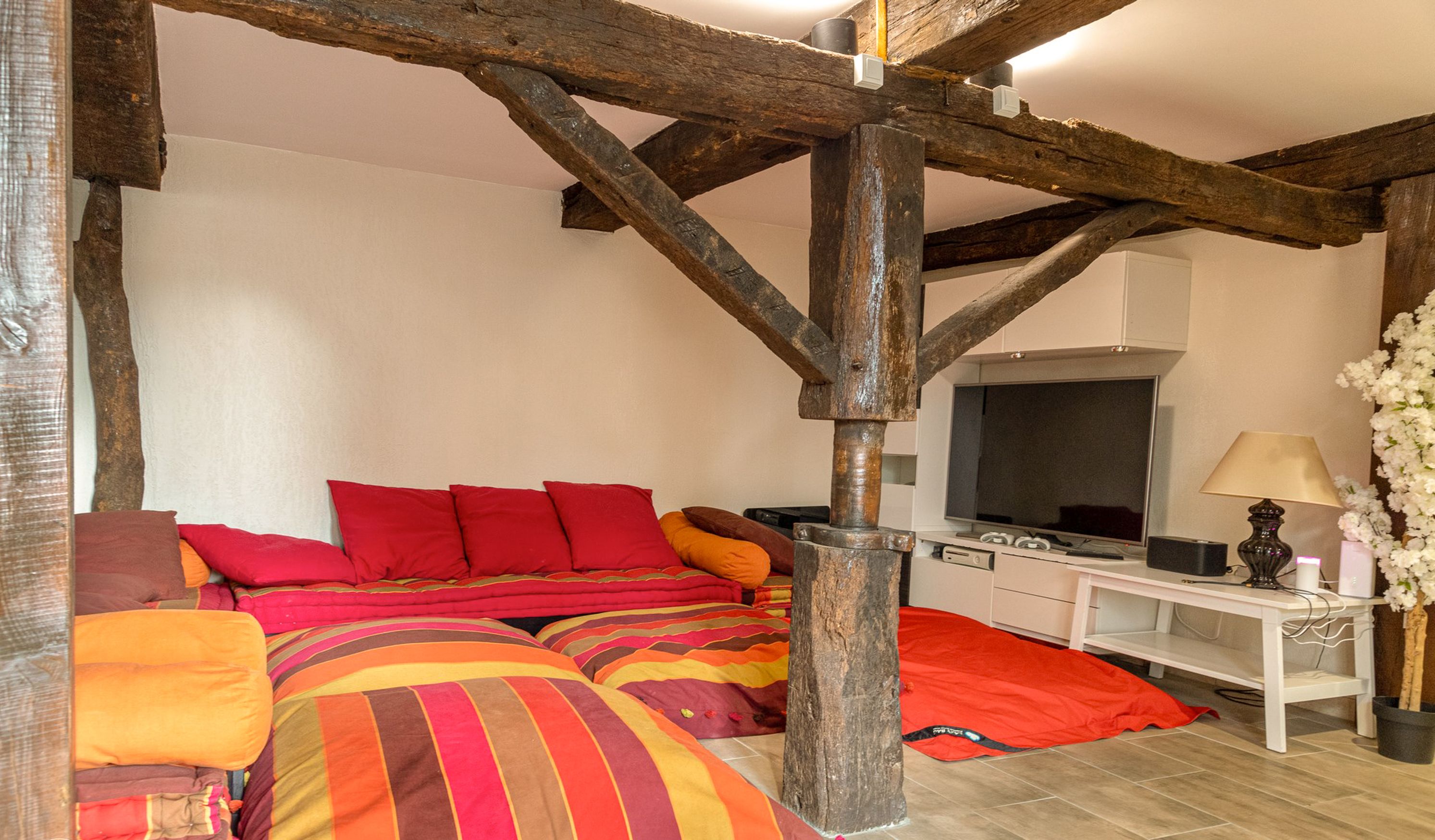 A bedroom with a sloped ceiling, wooden beams, a bed with colorful bedding, a television, and a bedside table with a lamp.