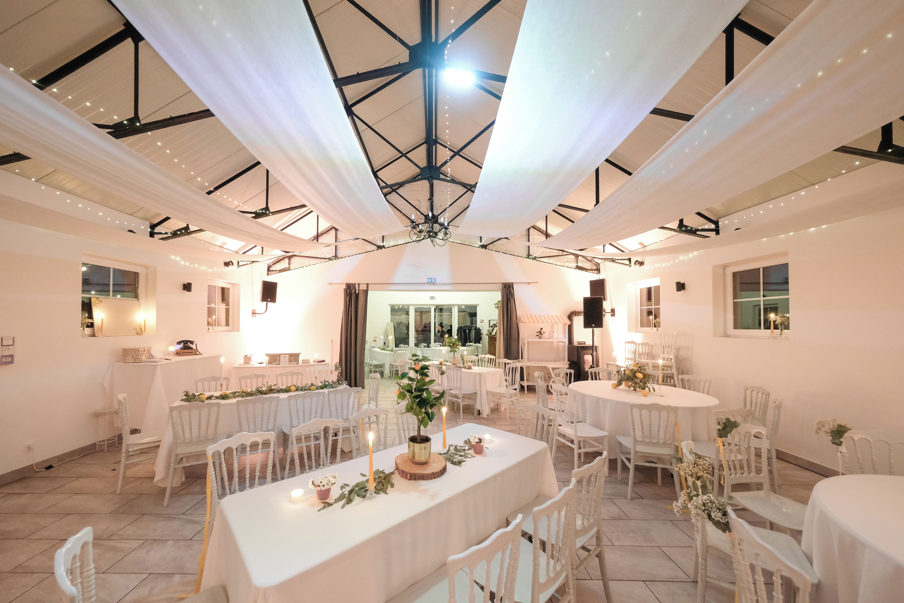 A spacious hall with multiple round tables and white chairs, featuring a high ceiling with a central light fixture.