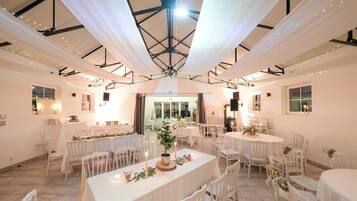 A spacious hall with multiple round tables and white chairs, featuring a high ceiling with a central light fixture.