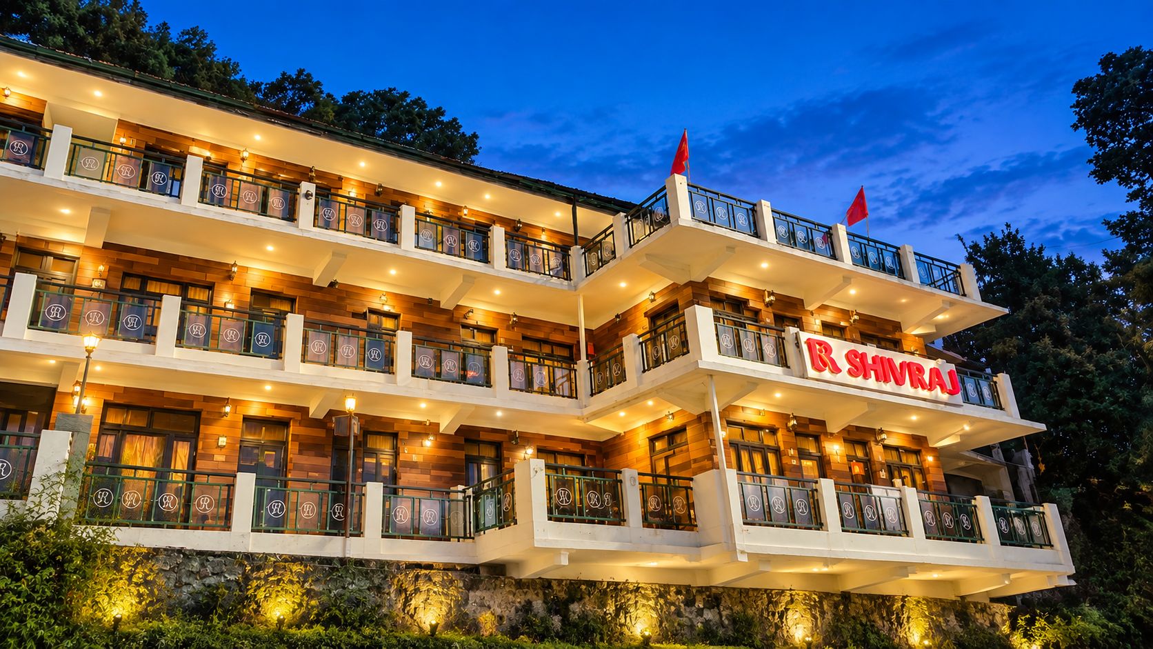 A multi-story hotel with balconies, a sign reading 'RS SHIVRAJ,' and warm lighting.