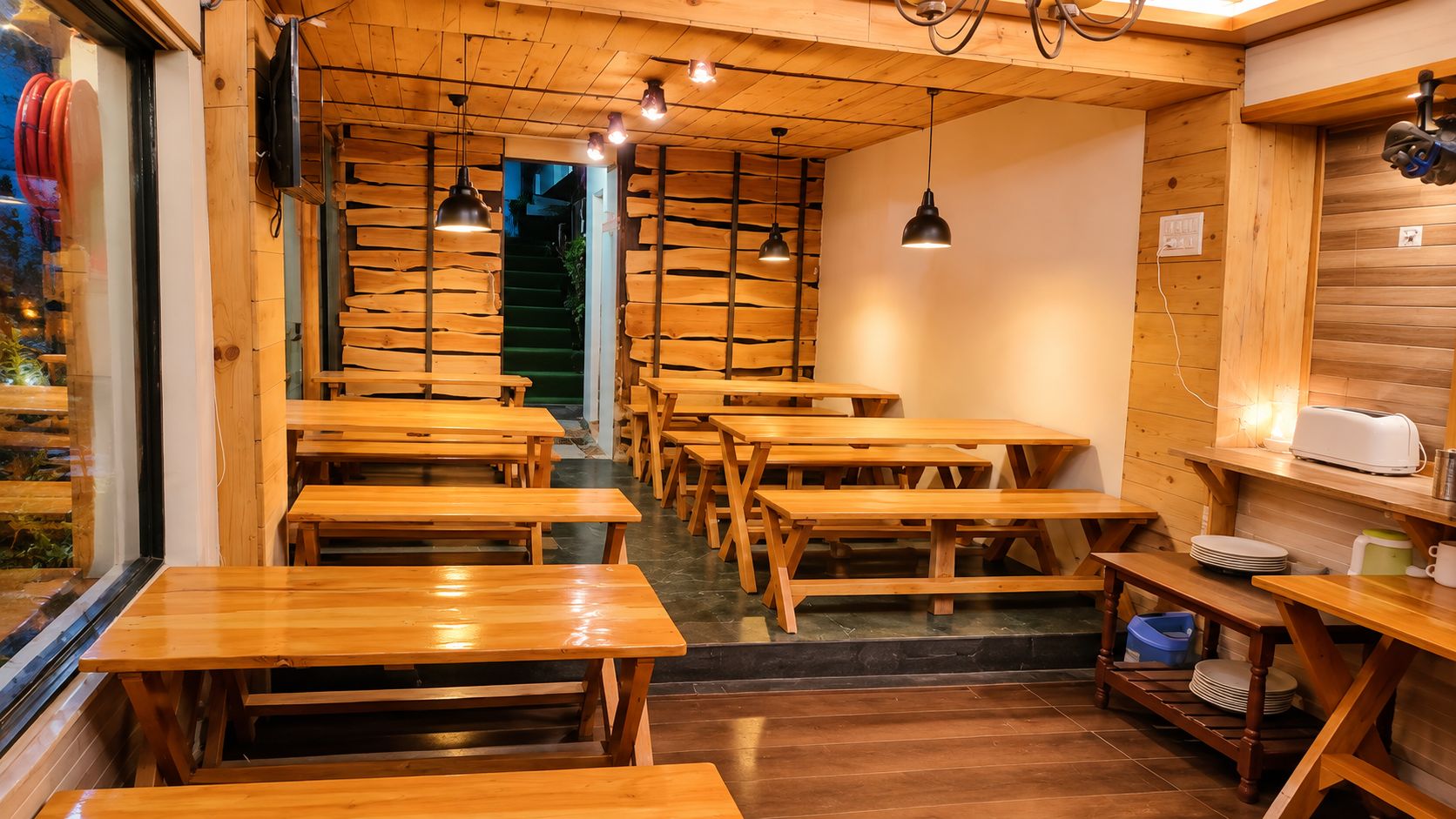 Interior de restaurante con mesas y bancos de madera, una escalera y lámparas colgantes.