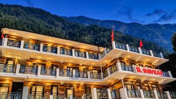 Un hotel de varios pisos con balcones, un cartel que dice 'R Shivraj' y un paisaje montañoso al fondo.
