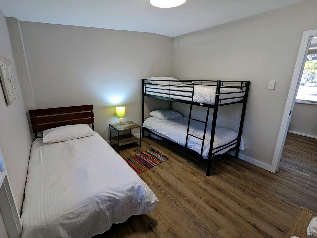 Una habitación con dos camas individuales, una litera, una mesita de noche con lámpara y suelo de madera.