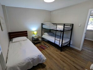 Ein Zimmer mit zwei Einzelbetten, einem Etagenbett, einem Nachttisch mit Lampe und Holzboden.