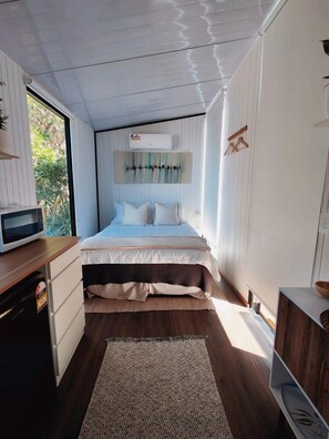 A bedroom with a bed, a bedside table, a microwave, and a window.
