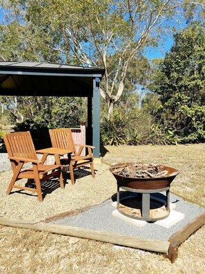 A covered outdoor seating area with two wooden chairs and a table, a fire pit, and a gazebo structure.