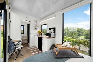 Habitació moderna amb llit, zona de menjador i petita cuina. Es veu verdura a l'exterior.