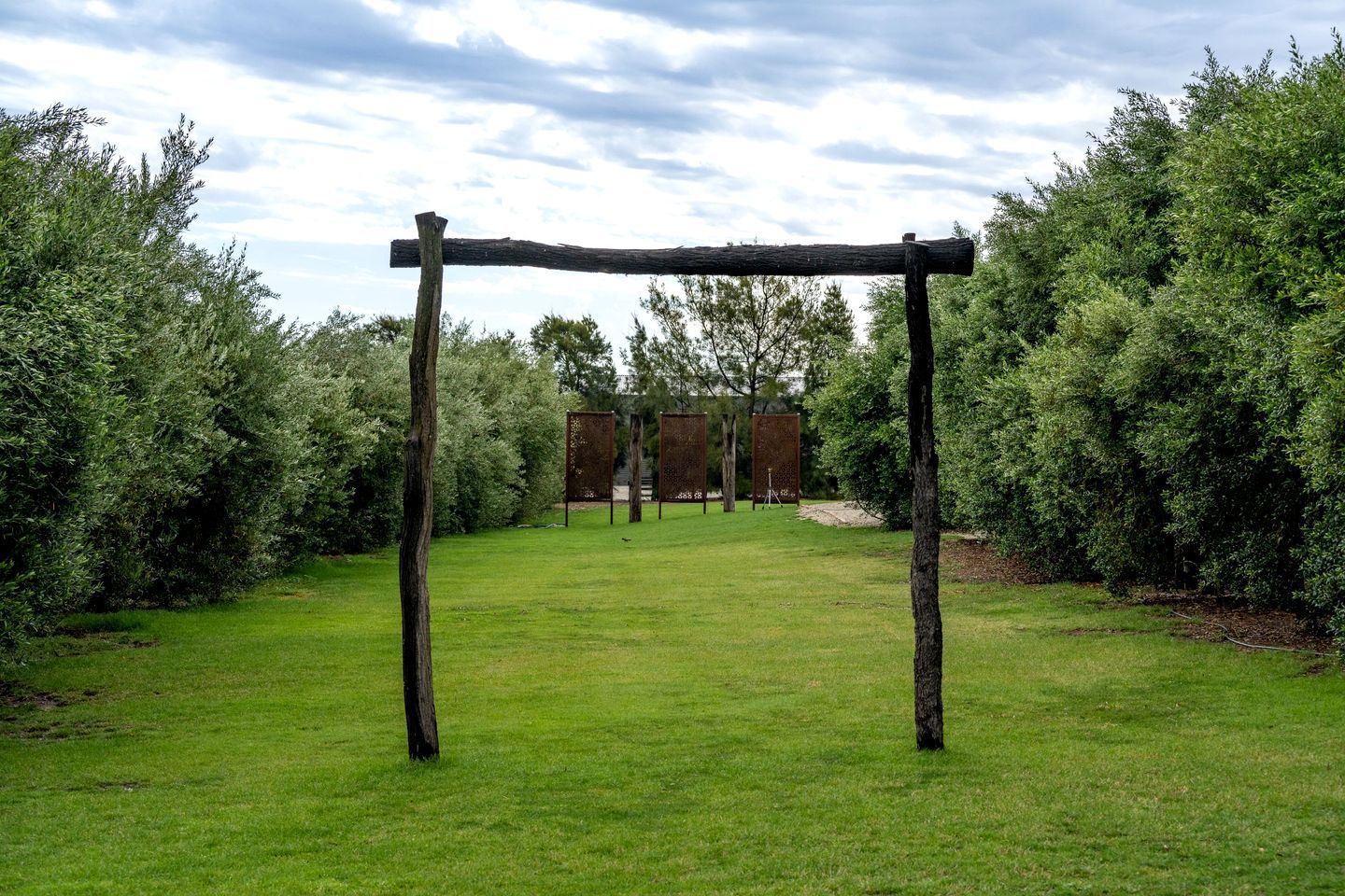 Une structure de pergola en bois, située dans une zone herbeuse, avec une végétation dense de chaque côté.
