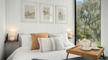 Een slaapkamer met een bed, kussens, een nachtkastje met een lamp, een raam met uitzicht op bomen en lijstwerken aan de muur.