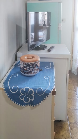 Ein kleines Zimmer mit einem Schreibtisch, einem Fernseher, einer Fernbedienung und einem dekorativen Behälter auf einer blauen Decke.