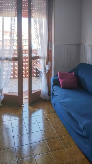 Ein Zimmer mit einem blauen Sofa, einem rosafarbenen Kissen, einem weißen Vorhang und einem Fliesenboden.