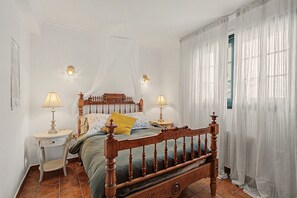 Ein Schlafzimmer mit Himmelbett, zwei Nachttischen mit Lampen, einem Fenster mit durchsichtigen Vorhängen und einem Fliesenboden.