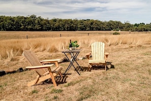 Zwei Adirondack-Stühle und ein kleiner Tisch mit einer Flasche und einem Glas in einer Wiese. Im Hintergrund ist ein Zaun und Bäume zu sehen.
