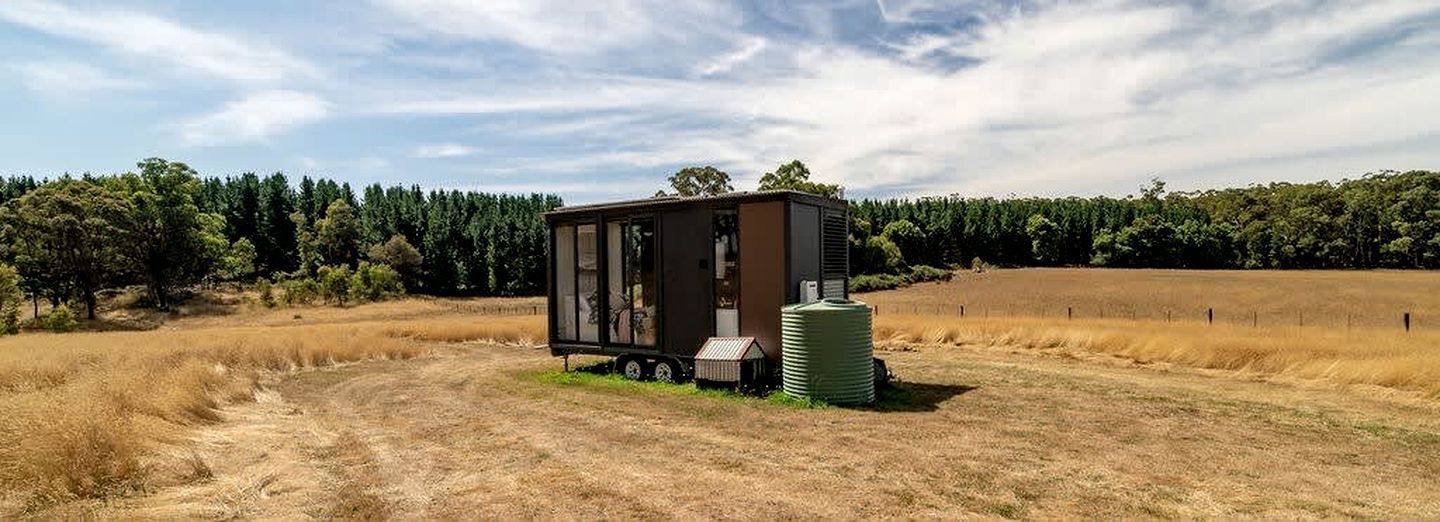 Una cabaña pequeña de un piso con fachada de metal, ubicada en una zona rural con un campo y árboles al fondo.