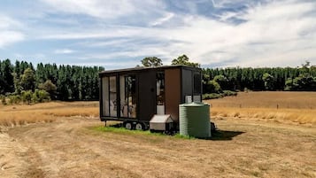 Una cabaña pequeña de un piso con fachada de metal, ubicada en una zona rural con un campo y árboles al fondo.